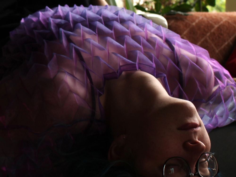 A photo of a person laying upside down on a chair. They are wearing a zig-zag pleated, sheer purple top, the light hitting the pleats and their neck which is craned to let their head hang towards the floor.