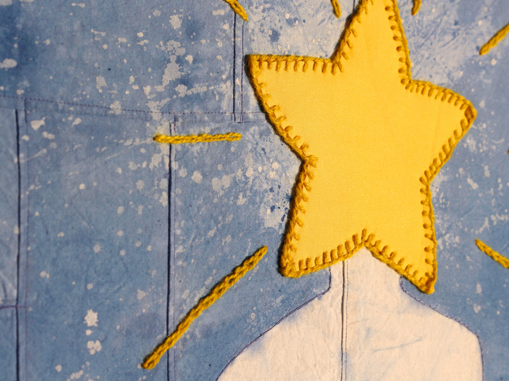 A close up photo of a yellow star applique on a patchwork blue fabric. The star is positioned to appear as the head of a body which has been resist dyed from the base fabric.