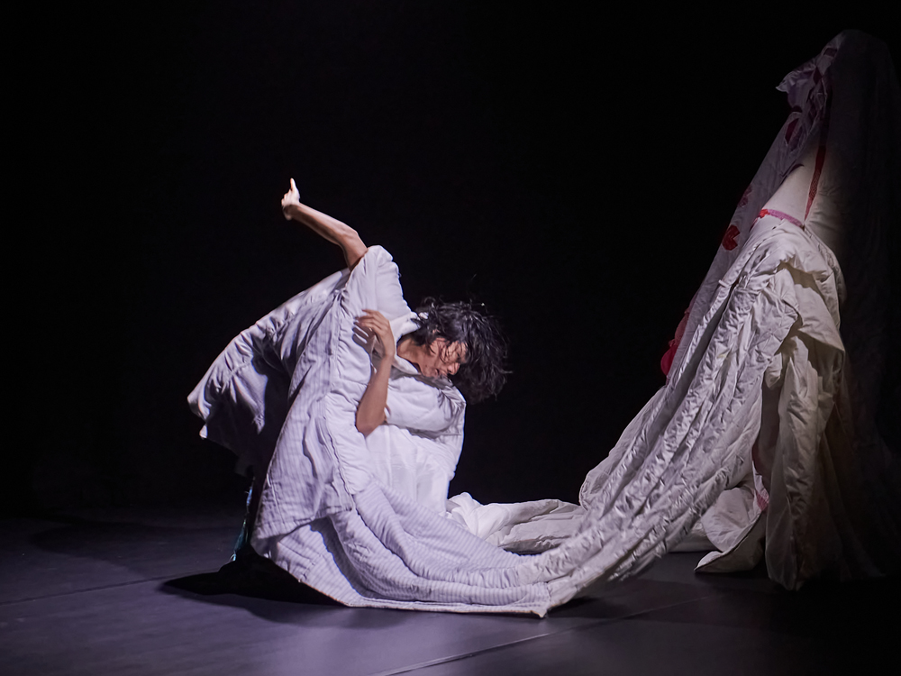A photo of a dancer in a dark room. The dancer is crouched low to the ground, mid-movement, with one arm to his chest and the other outstretched behind him. He wears a bright white, unshapely garment, made out of several sewn together duvets. The garment drops to the ground in front of him before continuing up into a pillar-like shape into the dark.