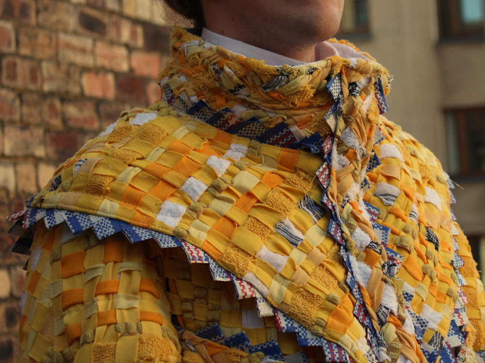 A photo of the upper body of a person wearing a yellow jacket. The garment has a large yoke and the pieces are made out of strips of yellow fabric woven together and stitched down.