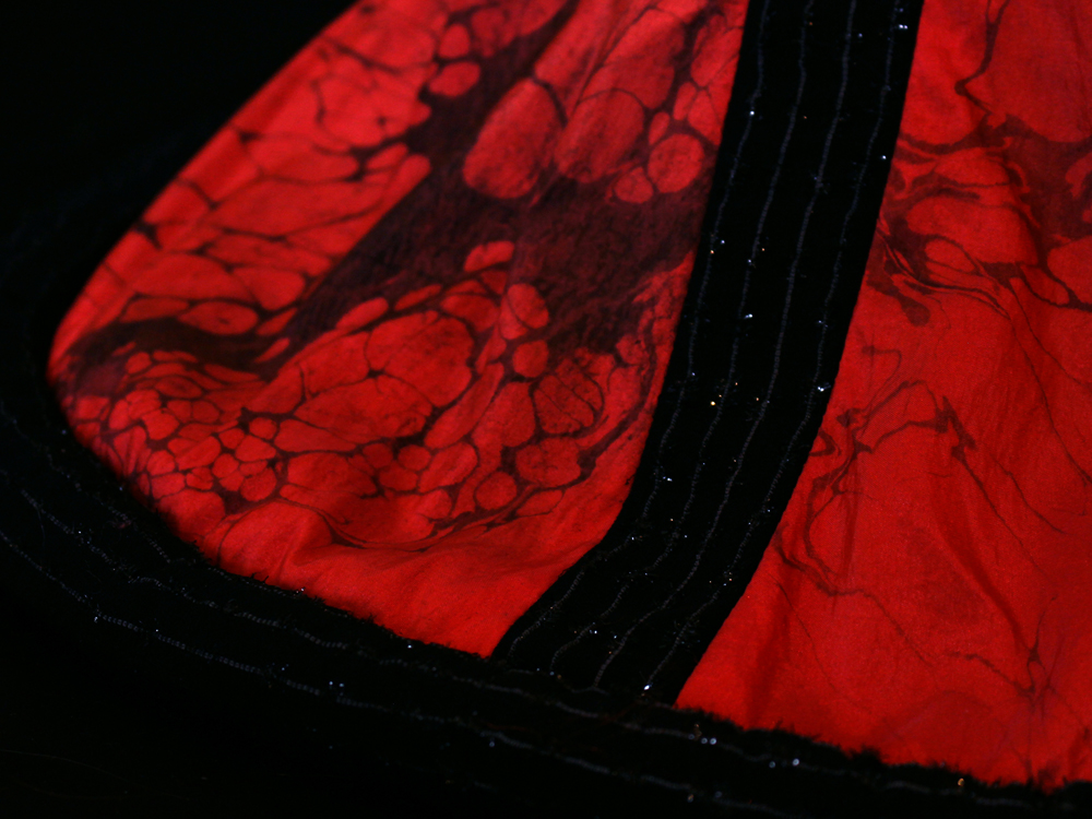 A photo of the hem of a garment. The garment is red with black marble-style surface decoration and black details. The hem is circular on the floor.