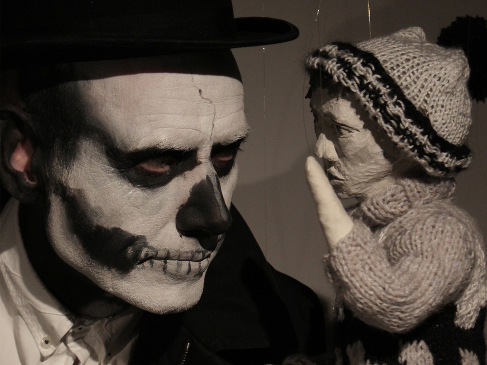 A photo of a puppeteer and a marionette. The puppeteer wears skull face paint and a black hat. The puppet has a carved wood face painted in greyscale and a knit sweater and hat, he has his hand raised as if to whisper to the puppeteer, who looks at him.