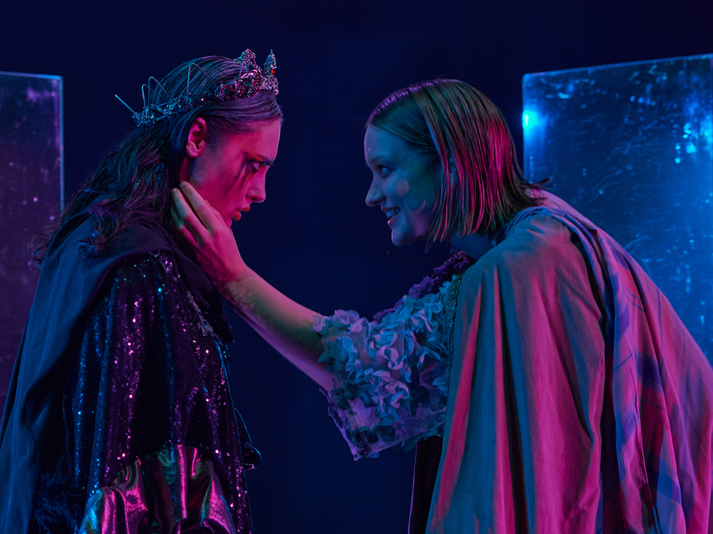 A photo of two actors facing each other. The left actor is dressed in grays and blacks with sparkling details, she wears a crown, her make-up is smeared down her face. The right actor wears a turquoise tulle top, a doublet and a cape, she holds the left one's face between her hands. They are lit in blue and pink.